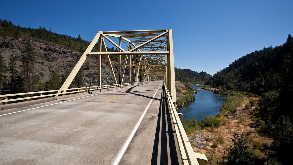 A concrete bridge with yellow railings spanning a river, surrounded by forested hills.