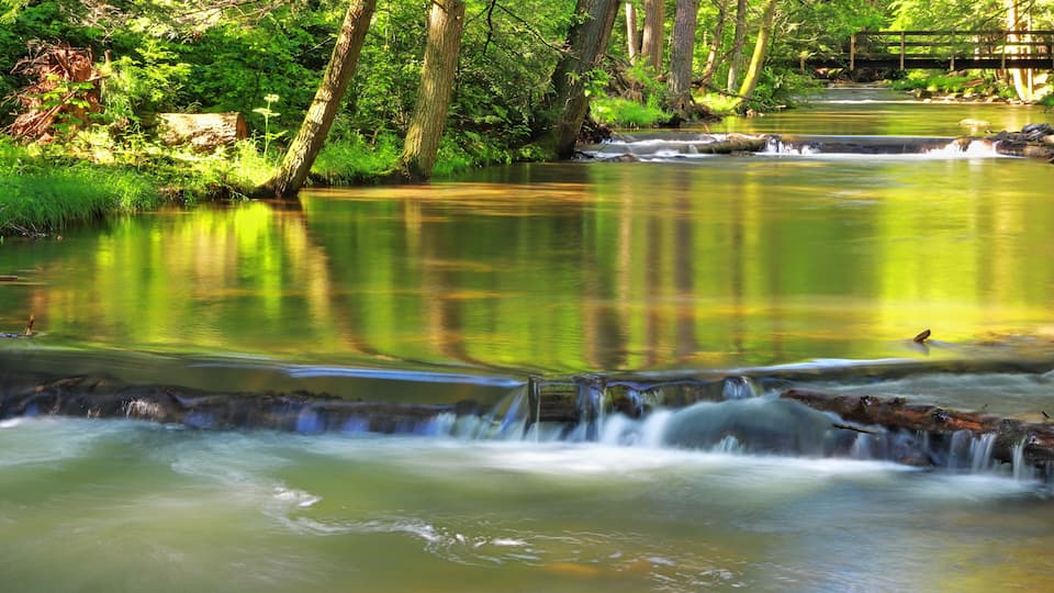 A calm river with a small waterfall, surrounded by lush greenery and trees.