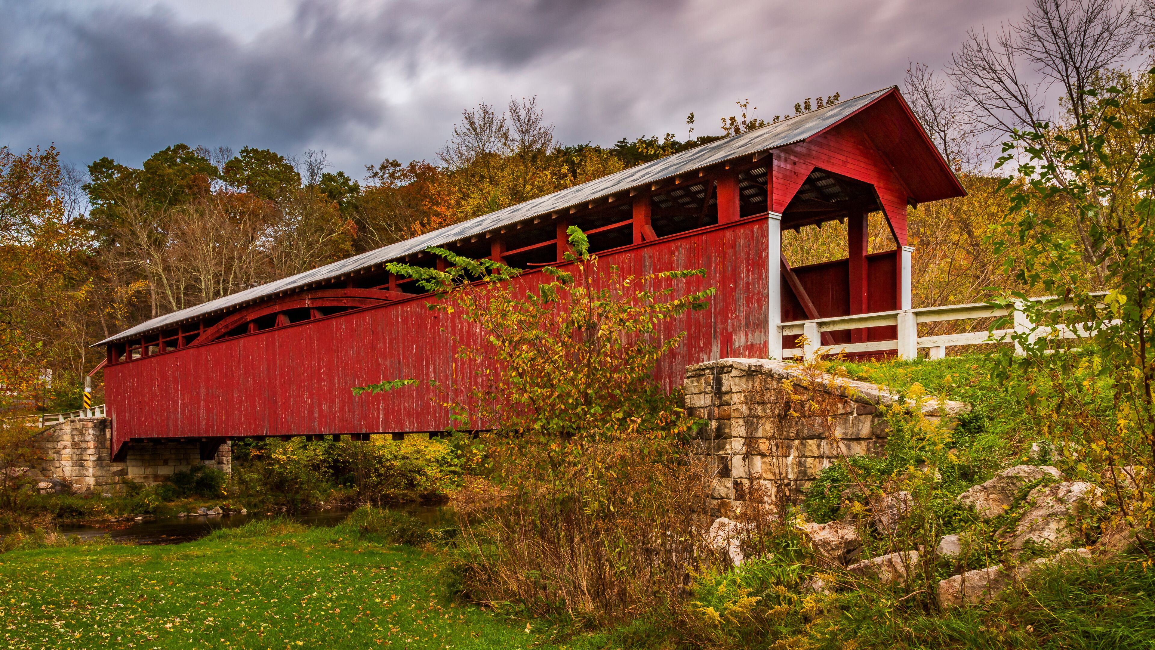 A red covered bridge with stone walls, surrounded by autumn foliage and a cloudy sky.