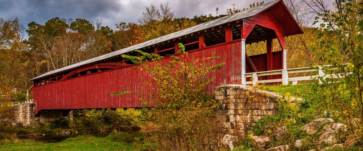 A red covered bridge with stone walls, surrounded by autumn foliage and a cloudy sky.