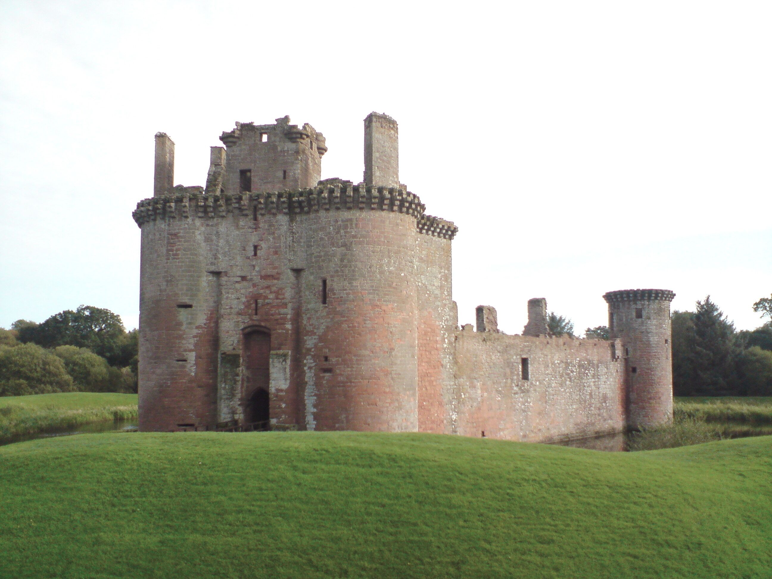 A large, historic castle with multiple towers and a central keep, surrounded by green fields.