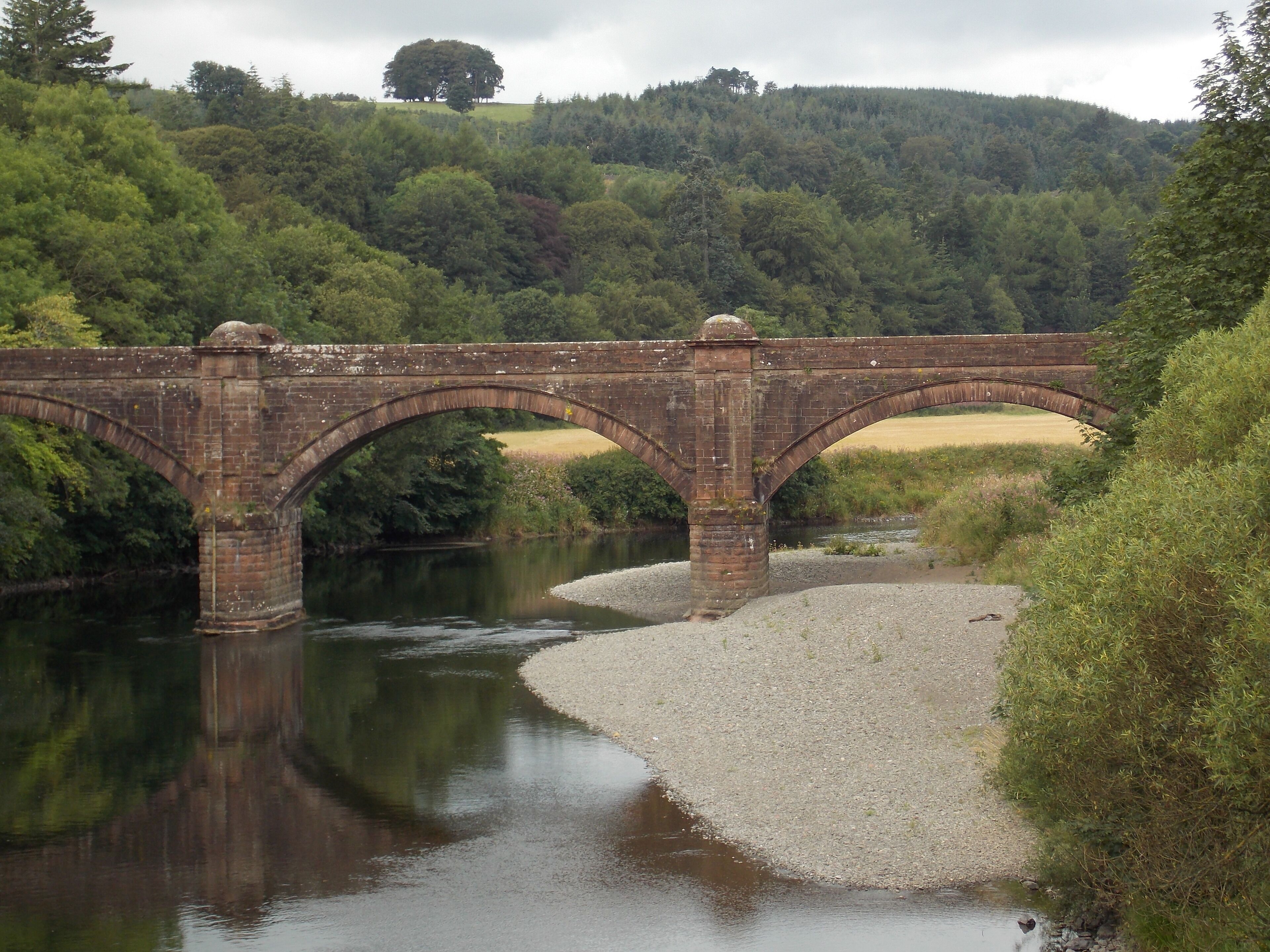 A stone bridge over a river with a forested hillside in the background.