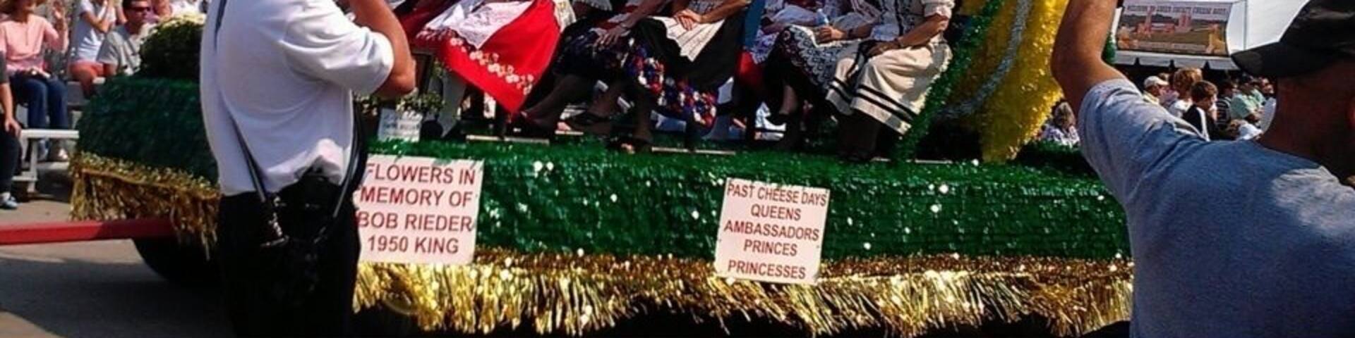 A parade float with a large yellow and green circular decoration and a sign reading "Flowers in Memory of Bob Riedler 1950 King."
