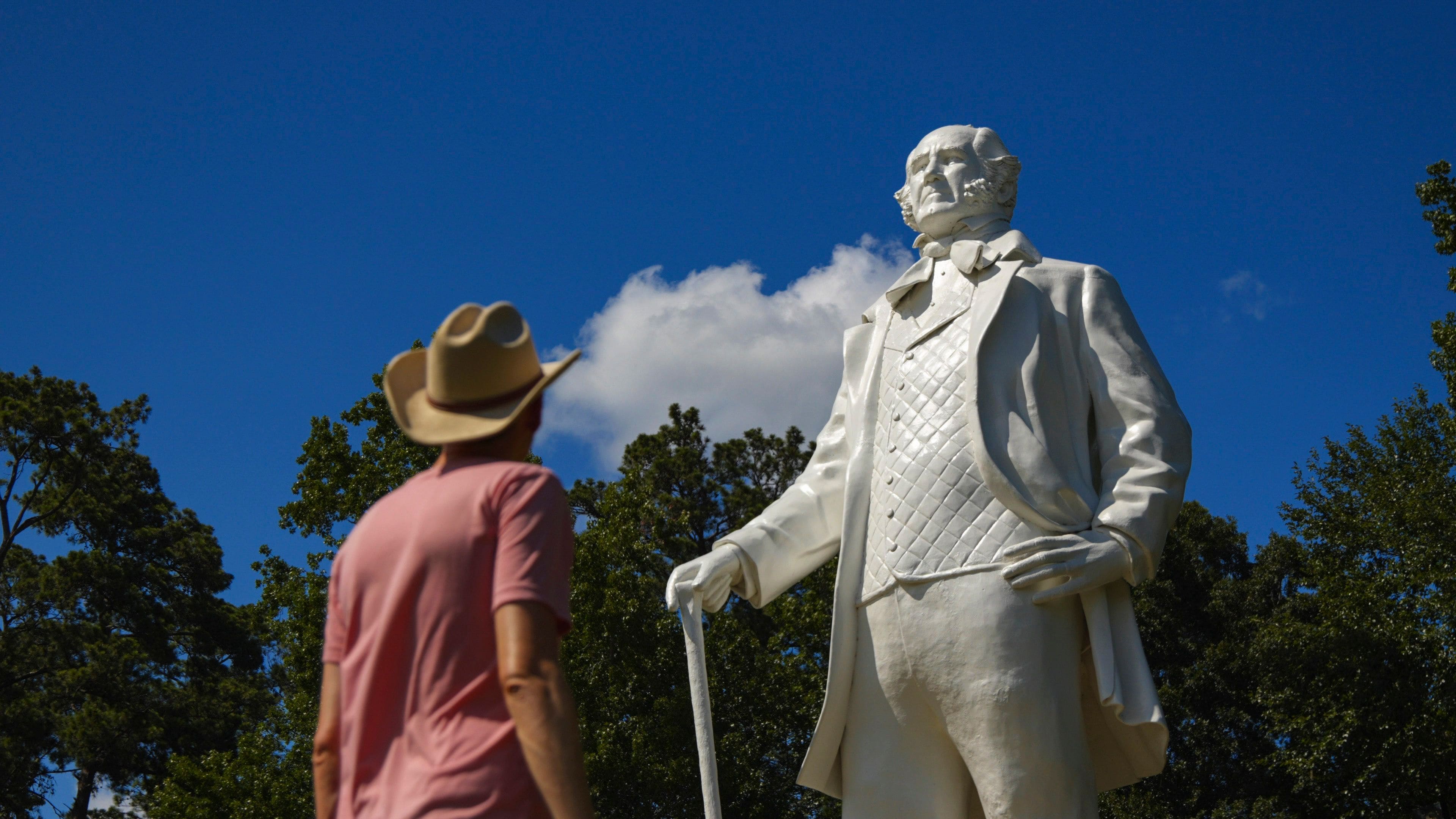 Eine Statue eines Mannes im Anzug und Hut, mit Stock, vor Bäumen.