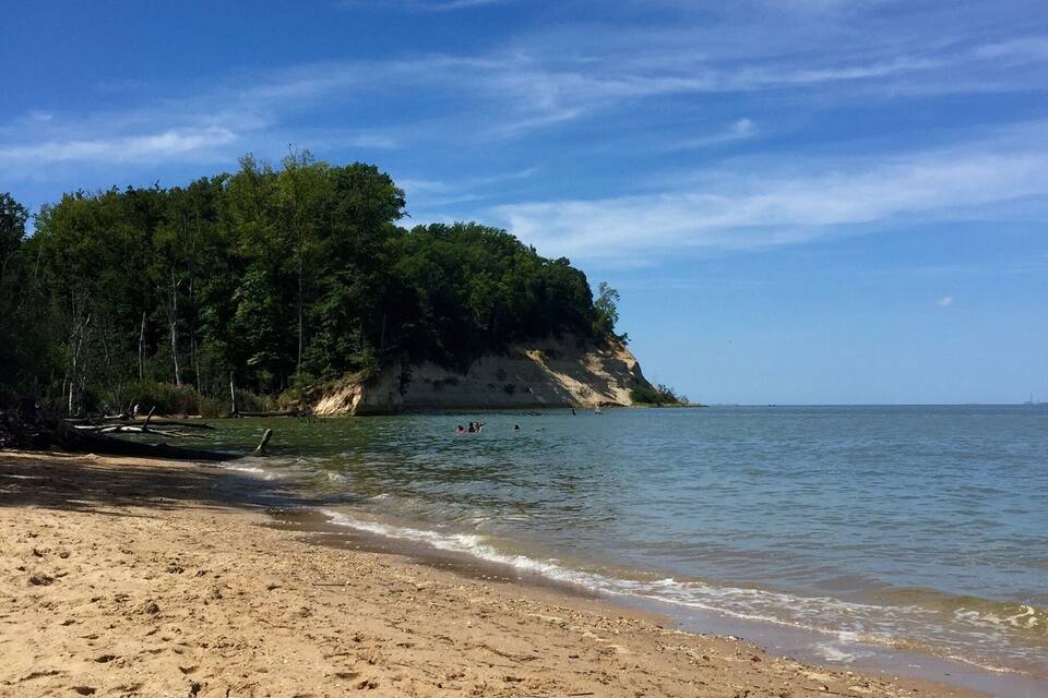 A sandy beach with a forested cliff and calm water.