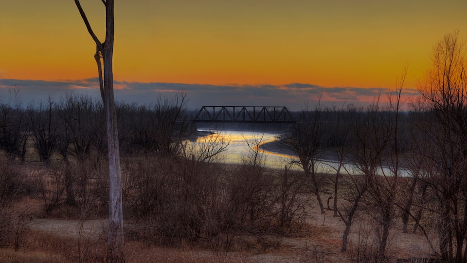 A sunset over a river with a bridge, surrounded by sparse trees and dry grass.