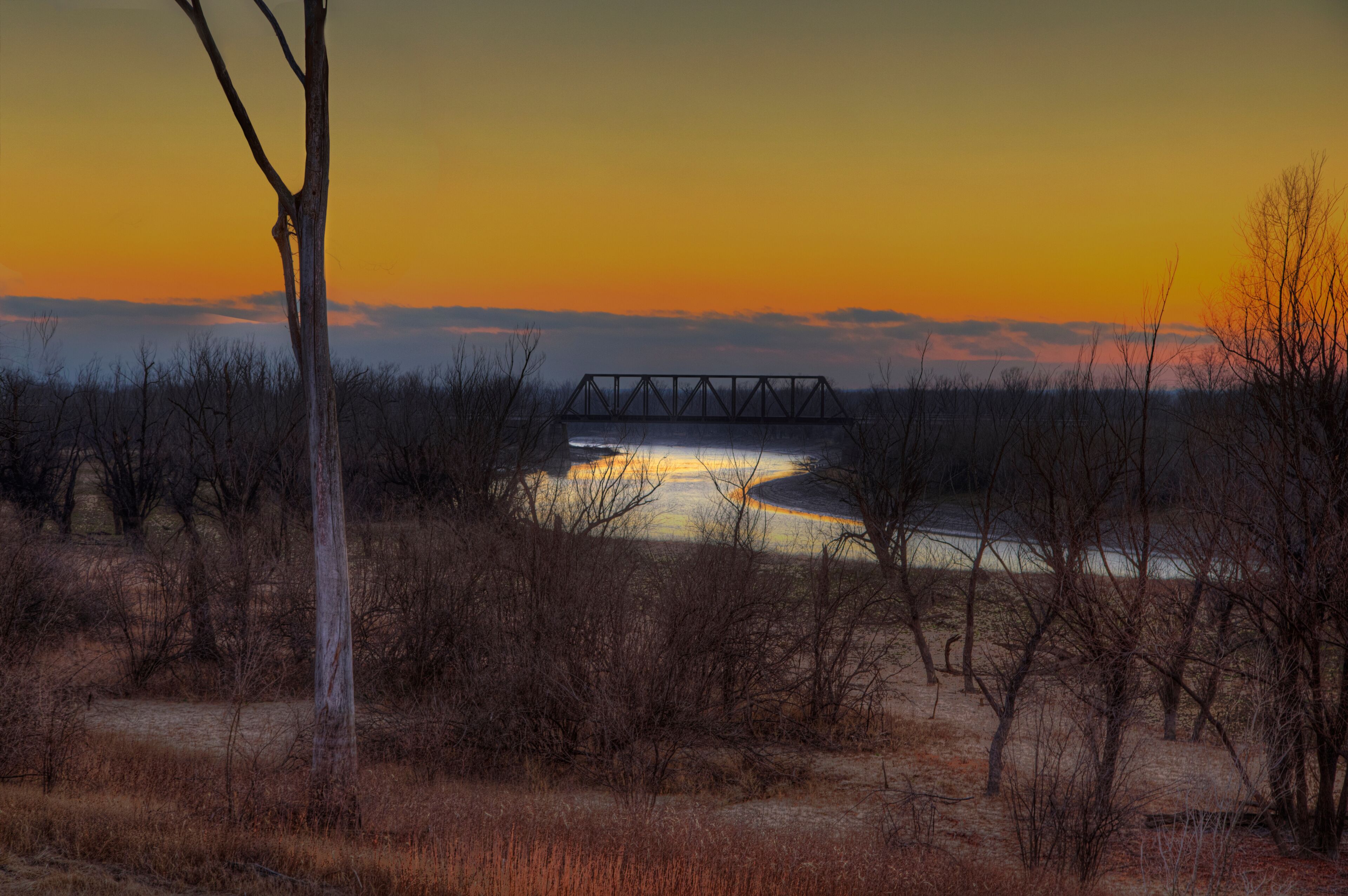 A sunset over a river with a bridge, surrounded by sparse trees and dry grass.