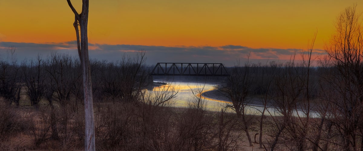 A sunset over a river with a bridge, surrounded by sparse trees and dry grass.