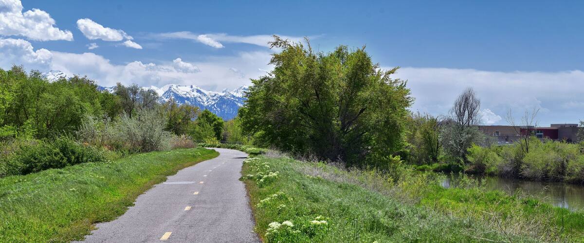 A paved road curving through a lush green landscape with a river on one side and a mountain range in the distance.