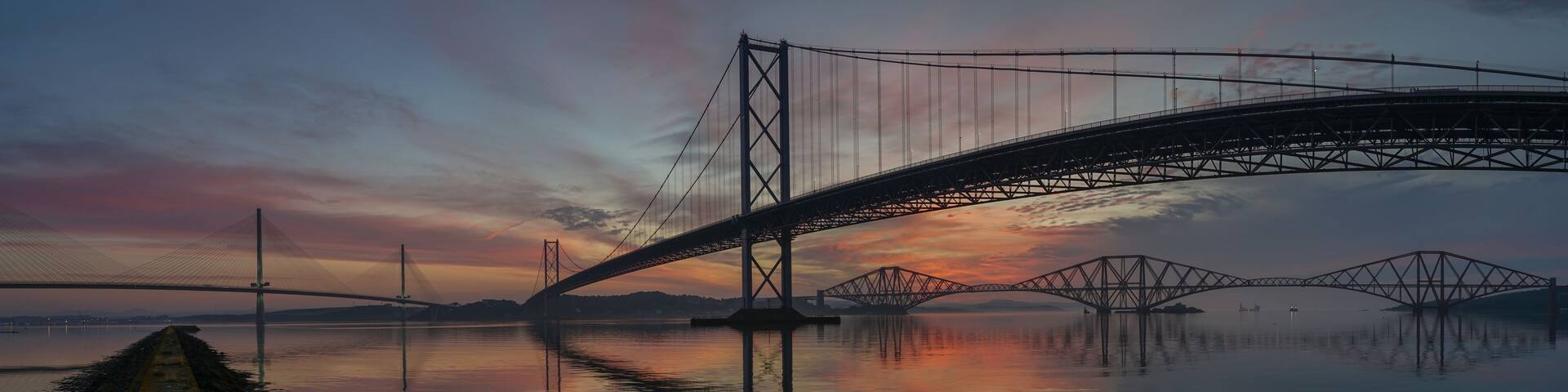 A suspension bridge over a calm body of water with a sunset in the background.