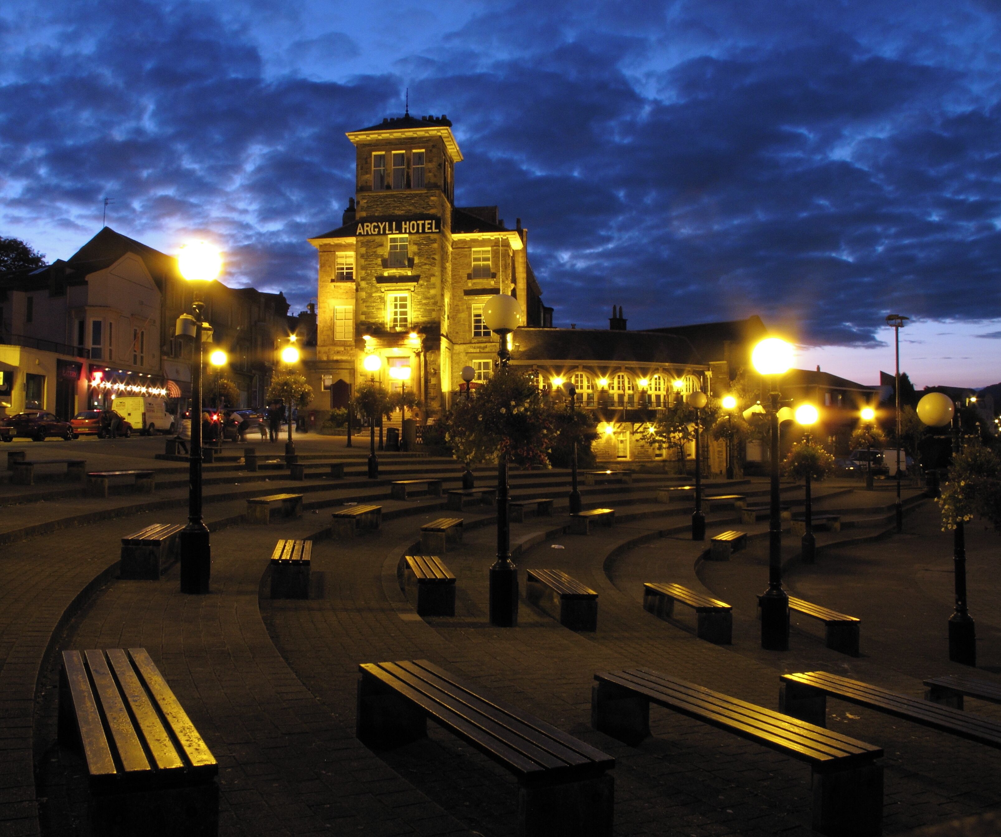 A historic building with a tower, surrounded by a well-lit public square with benches and street lamps.