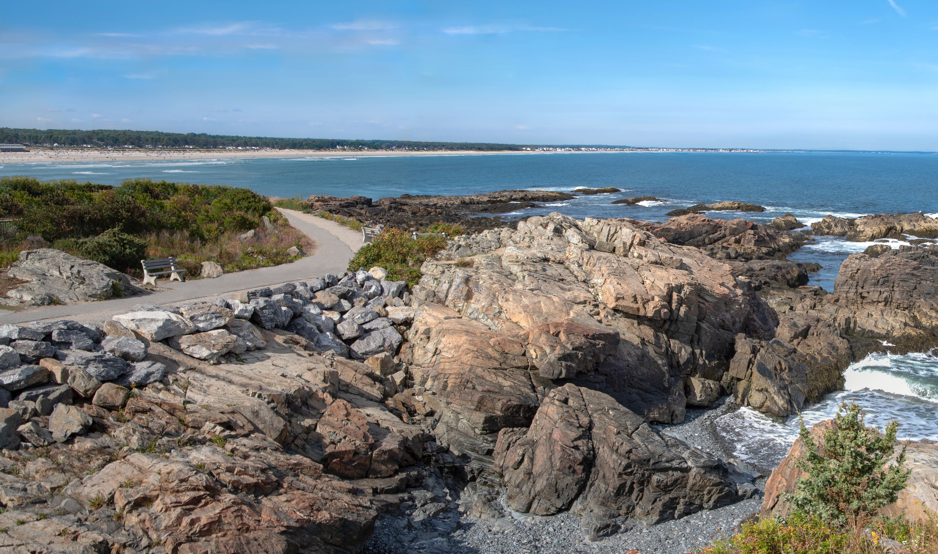 A rocky coastline with a winding path, a bench, and a view of the beach and ocean.