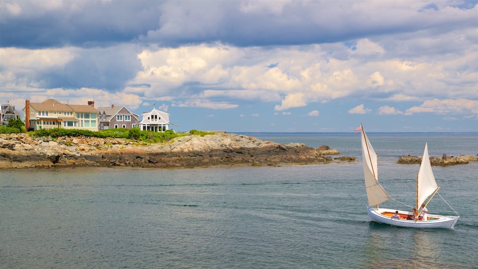 A sailboat on calm waters near a rocky shoreline with a row of houses.