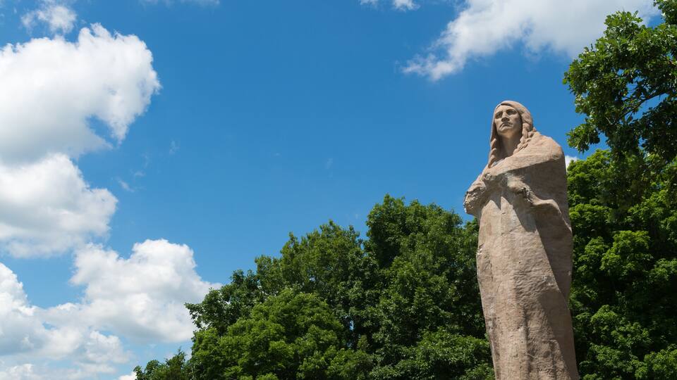 A large stone statue of a woman standing in a park with a clear blue sky and lush green trees in the background.