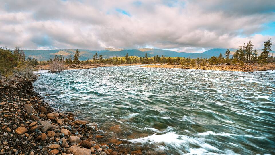 A river with a rocky shore, surrounded by trees and mountains under a cloudy sky.