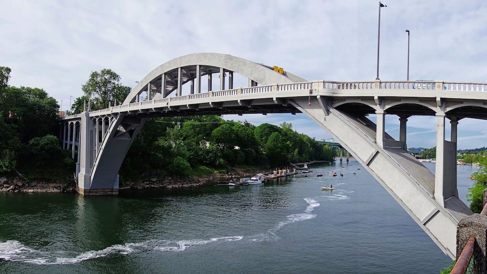 A large concrete bridge over a river with boats and greenery around.