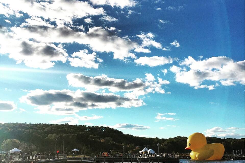 A beach with people, a large yellow duck sculpture, and a clear sky with clouds.
