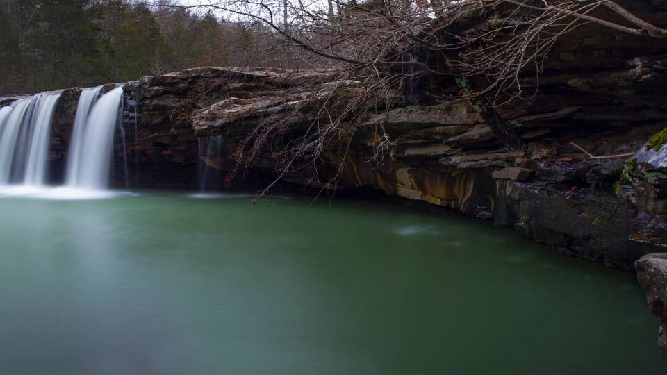 A waterfall cascading into a calm, greenish pool surrounded by rocky terrain and trees.