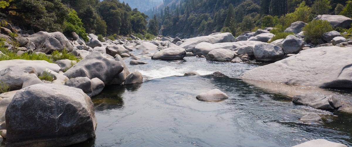 A river with large boulders, surrounded by dense forest and mountains.