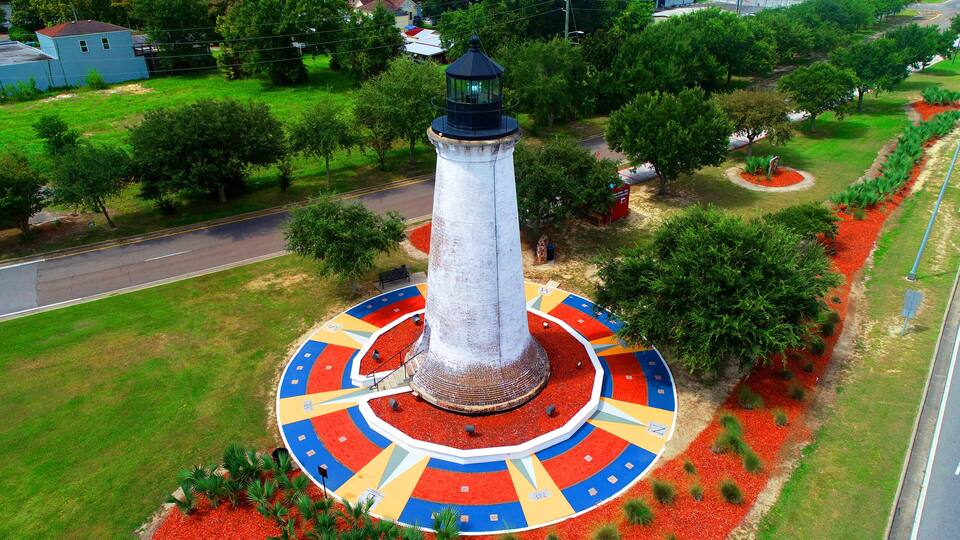 A lighthouse with a circular platform and colorful patterns, surrounded by trees and a road.