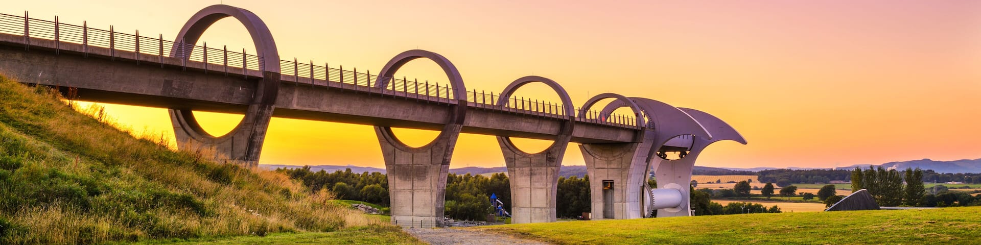 A bridge with circular arches over a grassy field during sunset.