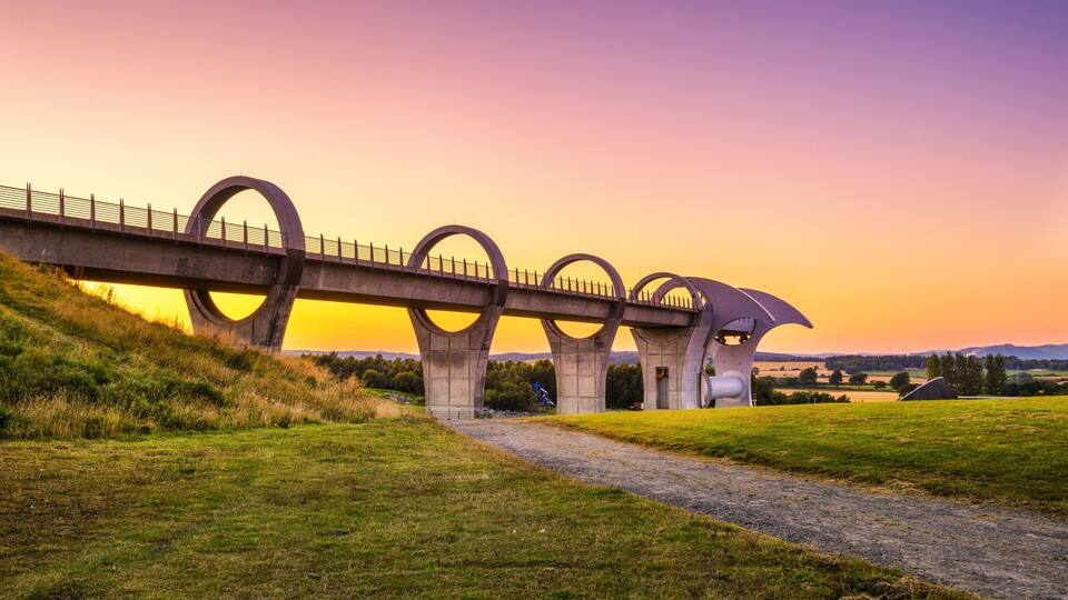 A bridge with circular arches over a grassy field during sunset.