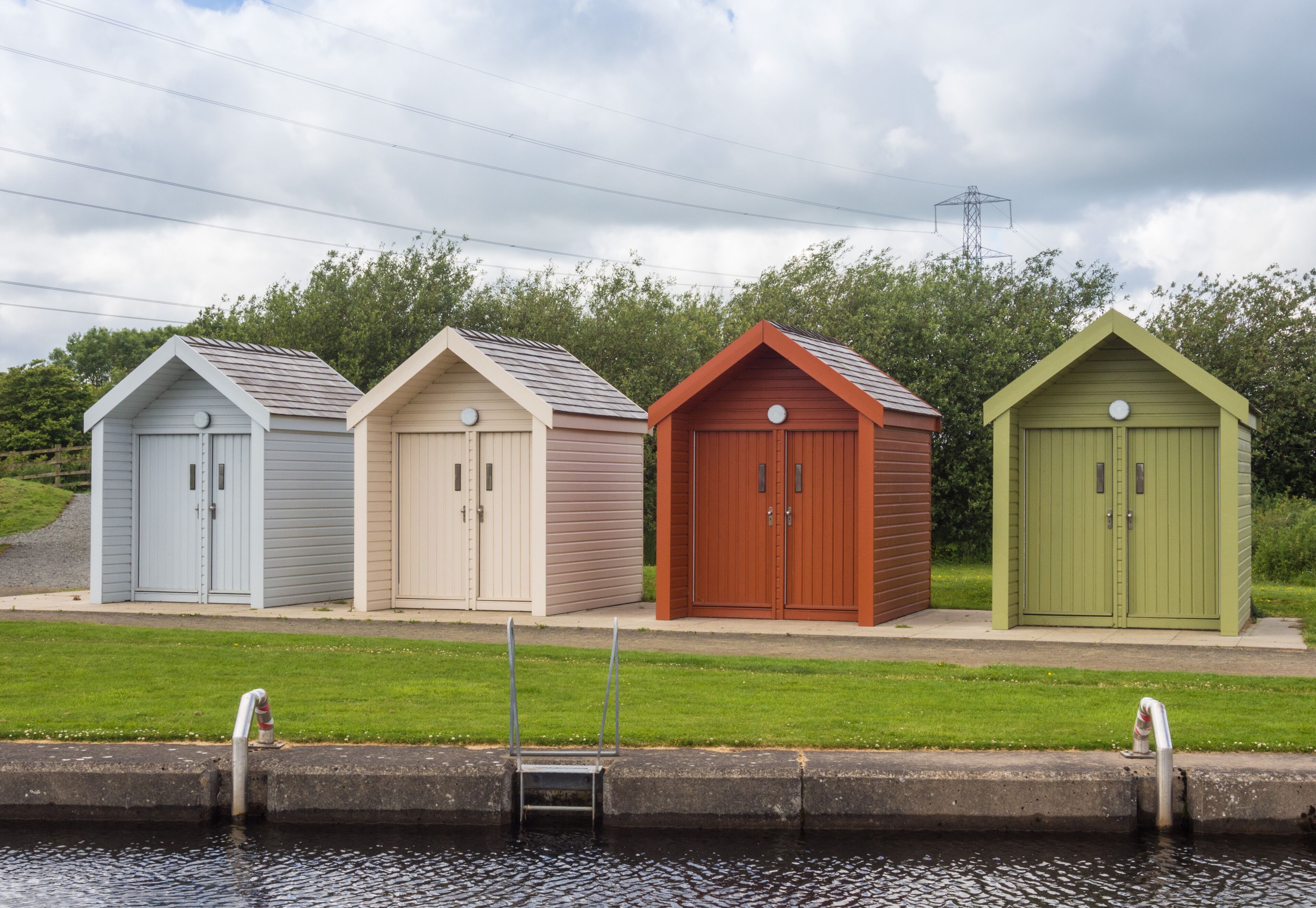 Four small sheds with different colors, a grassy area, and a body of water in the foreground.