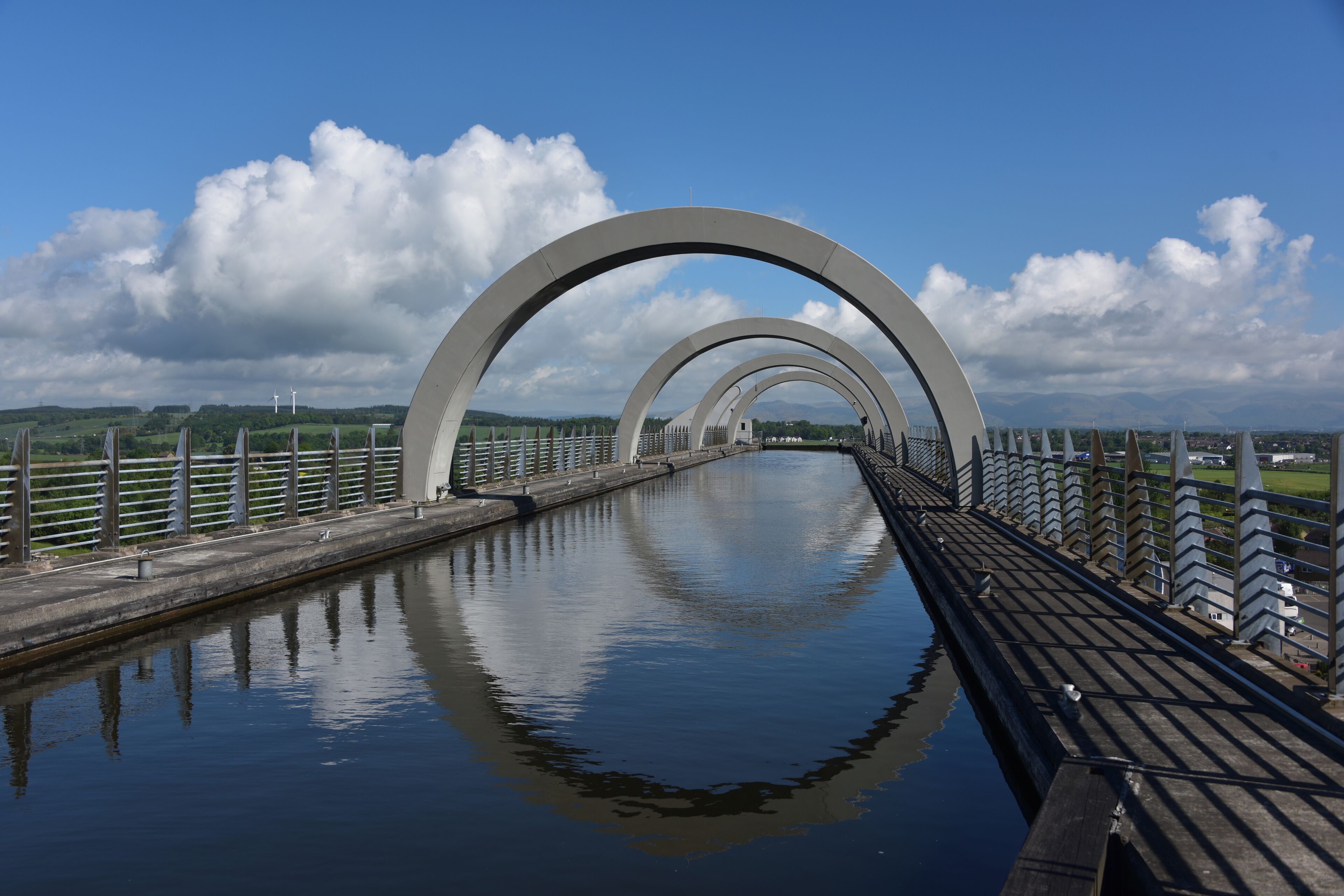 A bridge with large arches over a calm river, surrounded by greenery and distant wind turbines.