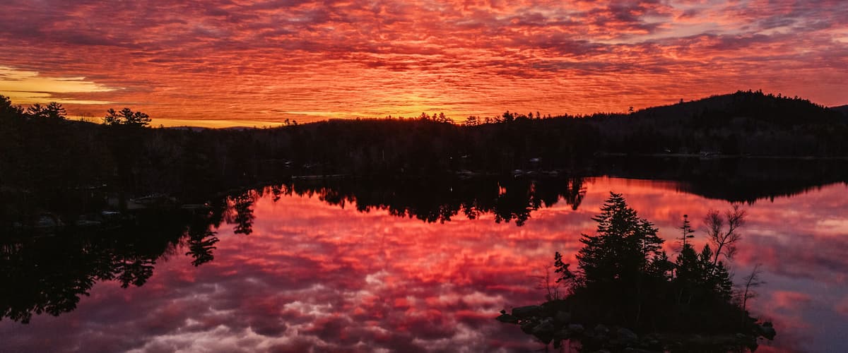 A sunset over a lake with red clouds reflected in the water, surrounded by trees and hills.