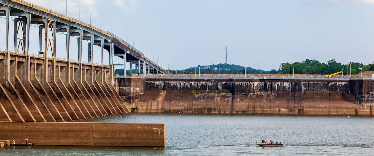A large dam with a bridge crossing over a river, boats on the water, and a distant hill with trees.