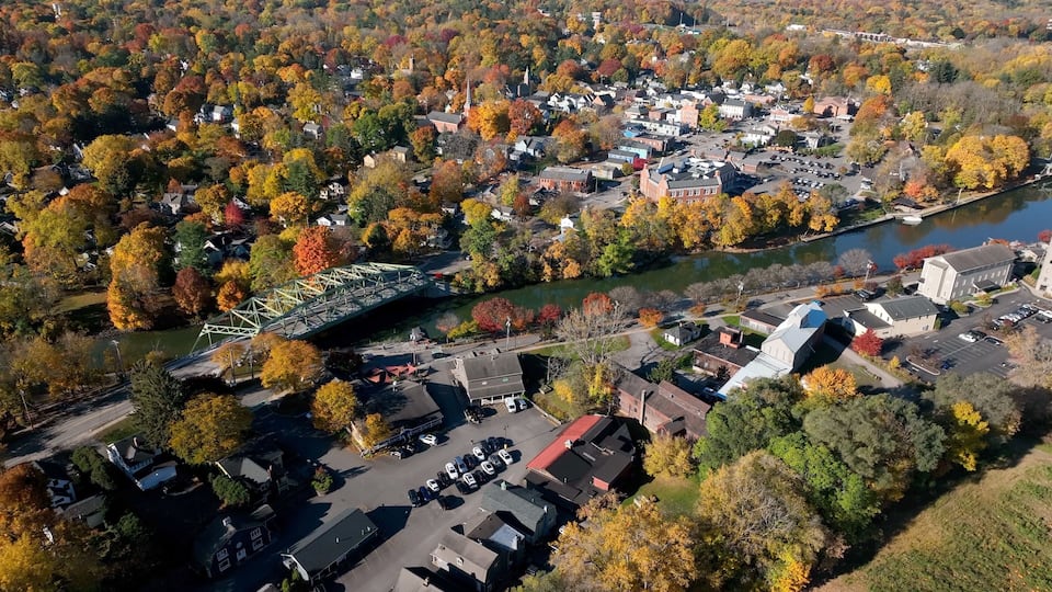 An aerial view of a town with a river, a bridge, and autumn-colored trees.