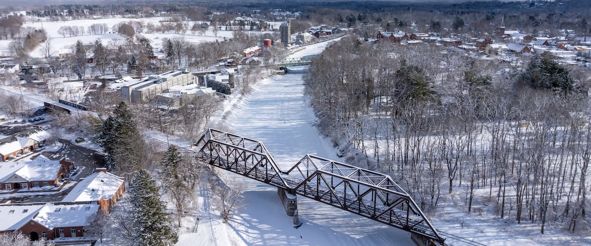 A snow-covered landscape with a bridge, buildings, and a river.