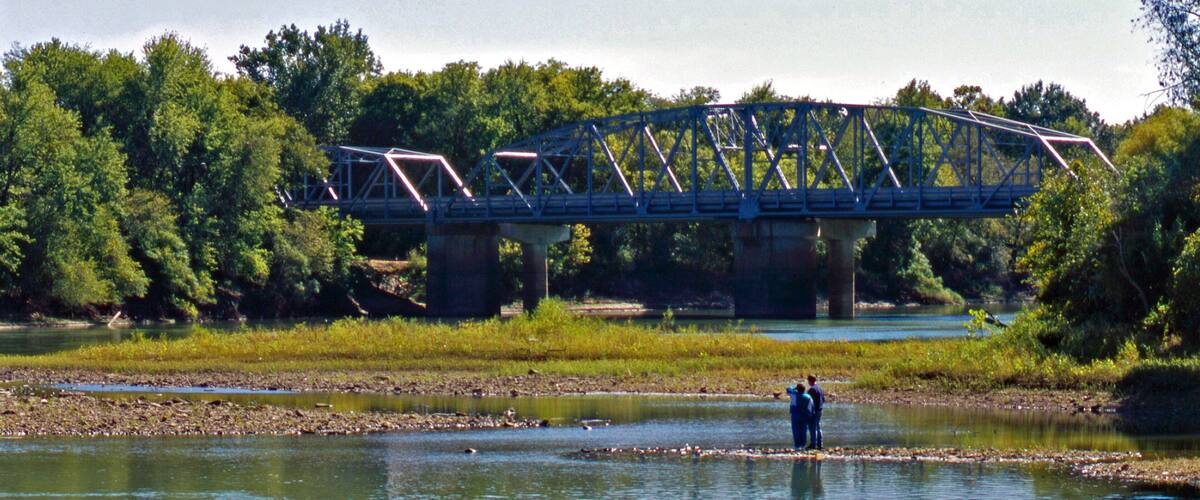 A steel bridge over a river with people standing on the riverbank.