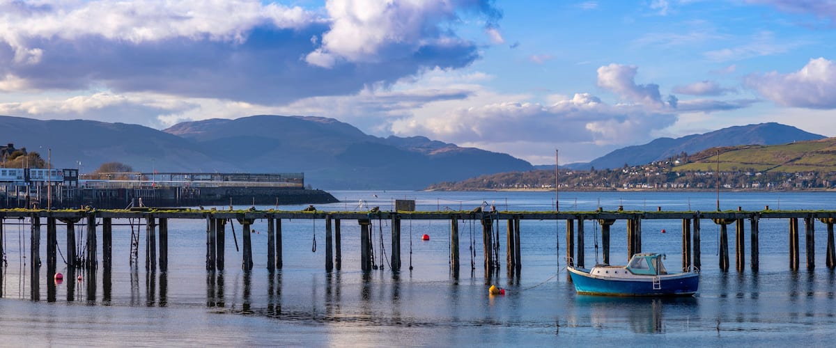 A coastal scene with a pier extending into calm waters, a small boat moored at the end, and mountains in the background.