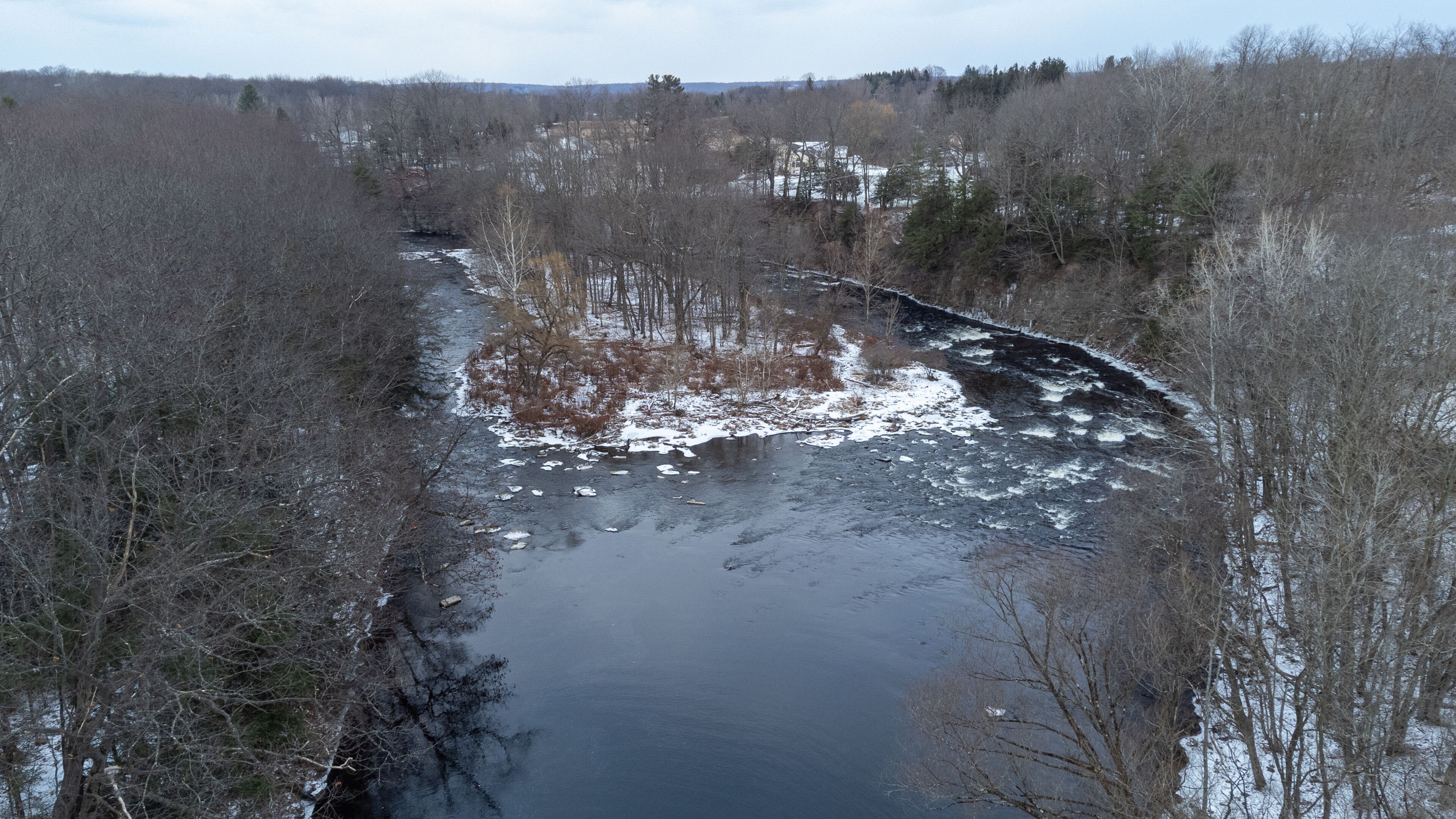 A river flowing through a forested area with snow patches on the ground.