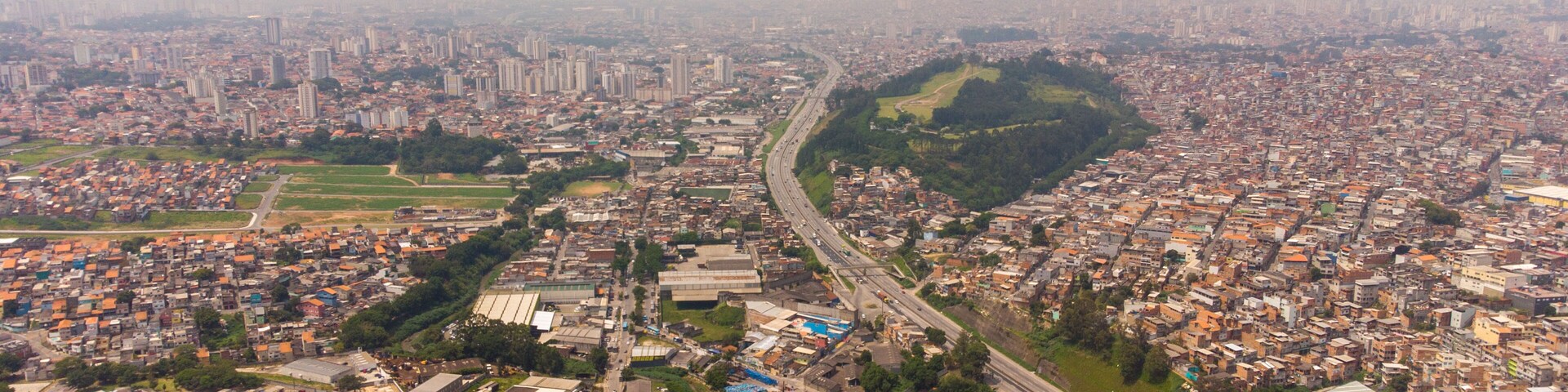 Uma área urbana densamente povoada, com uma mistura de edifícios residenciais e comerciais, um parque central e uma autoestrada atravessando a cidade.