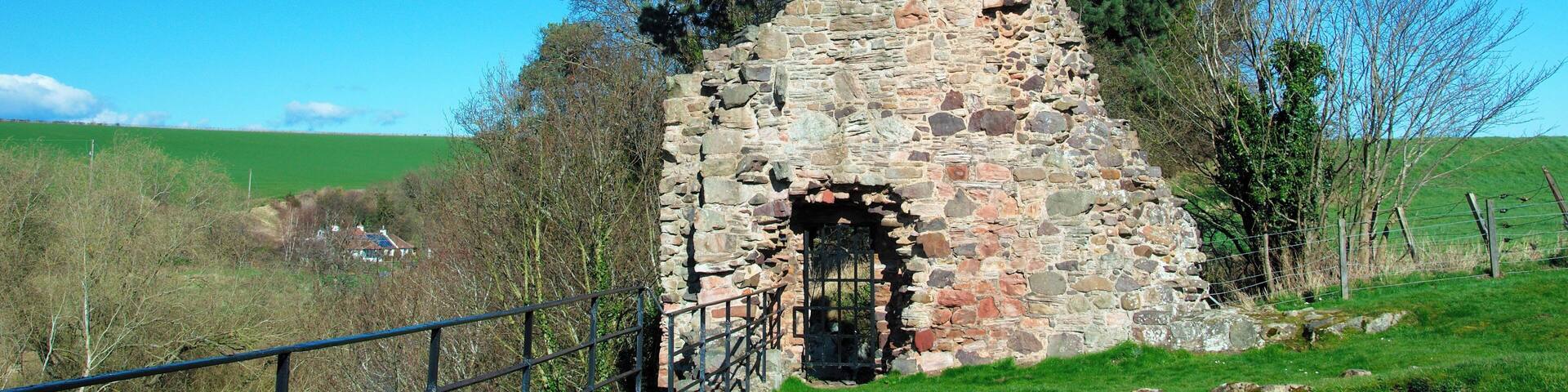 A stone ruin with a wooden door, surrounded by trees and a grassy field.