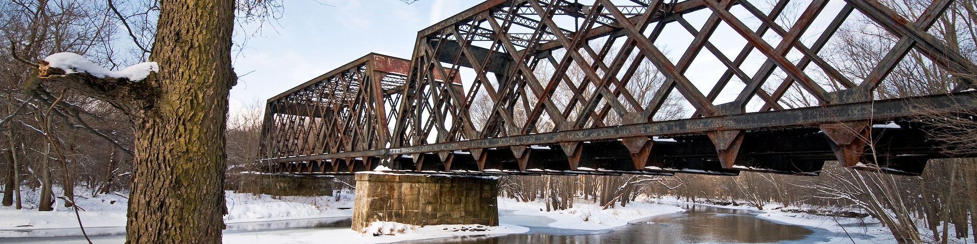 Un puente metálico oxidado sobre un río helado, con nieve en el suelo y árboles al fondo.