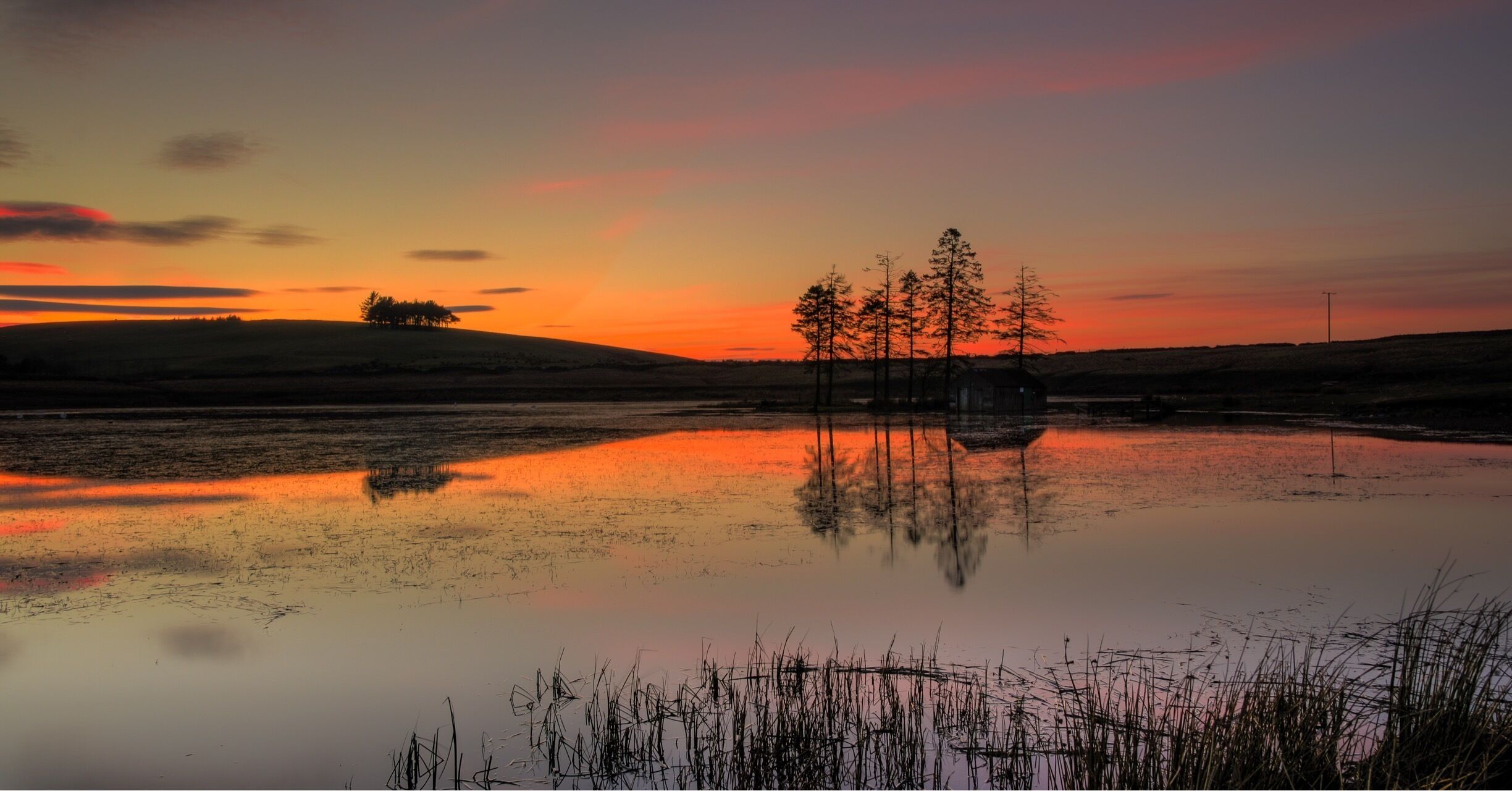 A sunset over a calm lake with trees reflecting the sky, a distant hill, and a small structure.