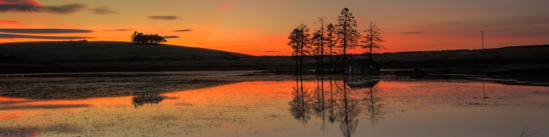 A sunset over a calm lake with trees reflecting the sky, a distant hill, and a small structure.