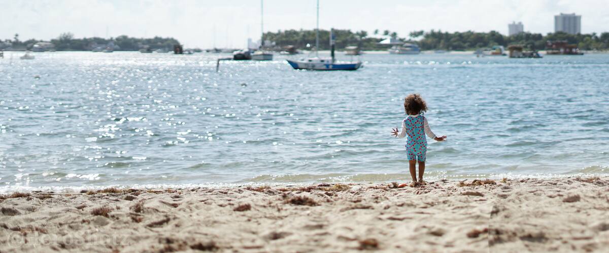 A child standing on a sandy beach, looking at the ocean with boats in the distance.