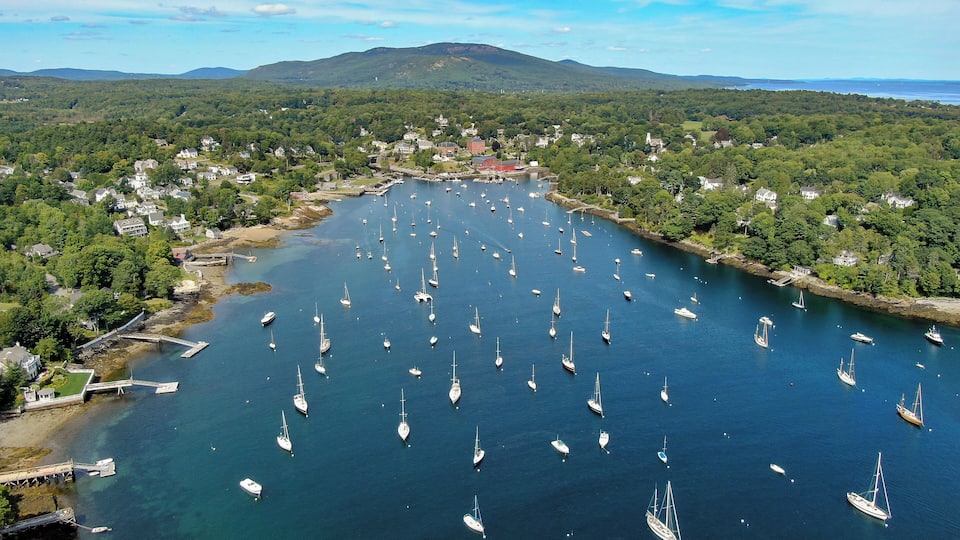 A coastal town with numerous sailboats anchored in a bay surrounded by lush greenery and hills.