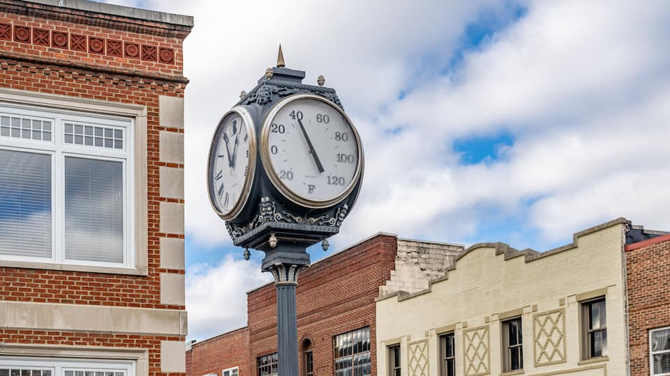 A clock tower with two faces, set against a backdrop of brick buildings and a partly cloudy sky.