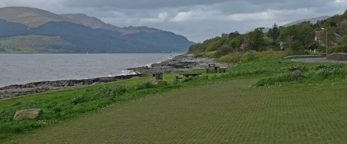 A coastal landscape with a grassy area, a rocky shoreline, and mountains in the background.