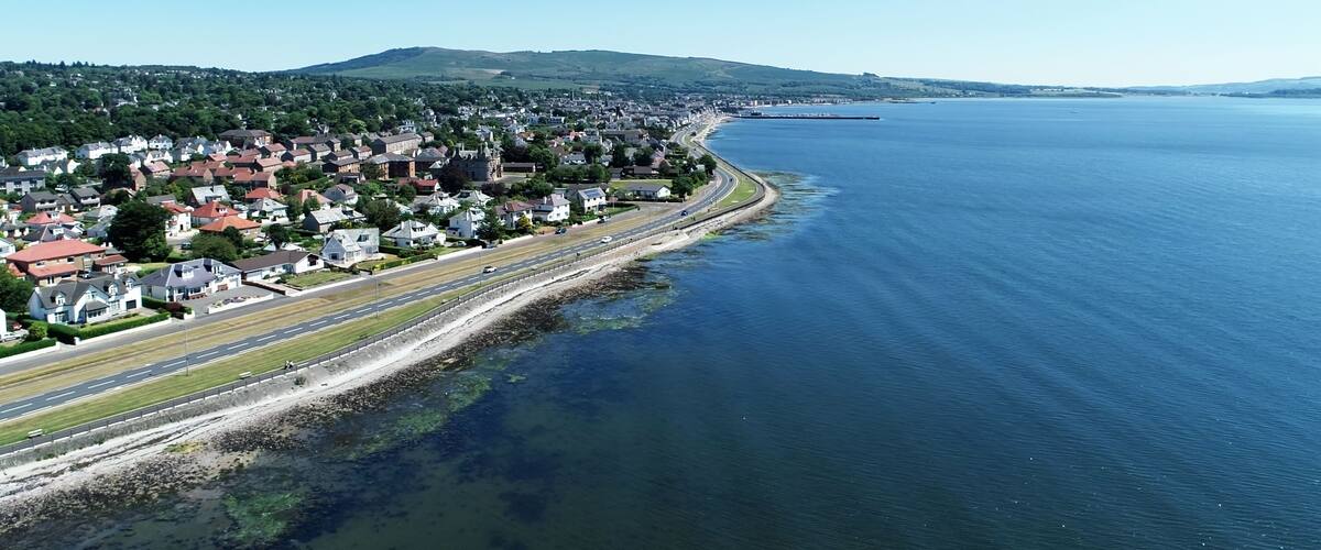A coastal town with a road running parallel to the water, houses, and a distant mountain range.