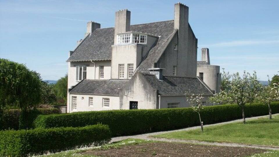 A large, white, two-story house with a grey roof, surrounded by a well-maintained garden and hedges.