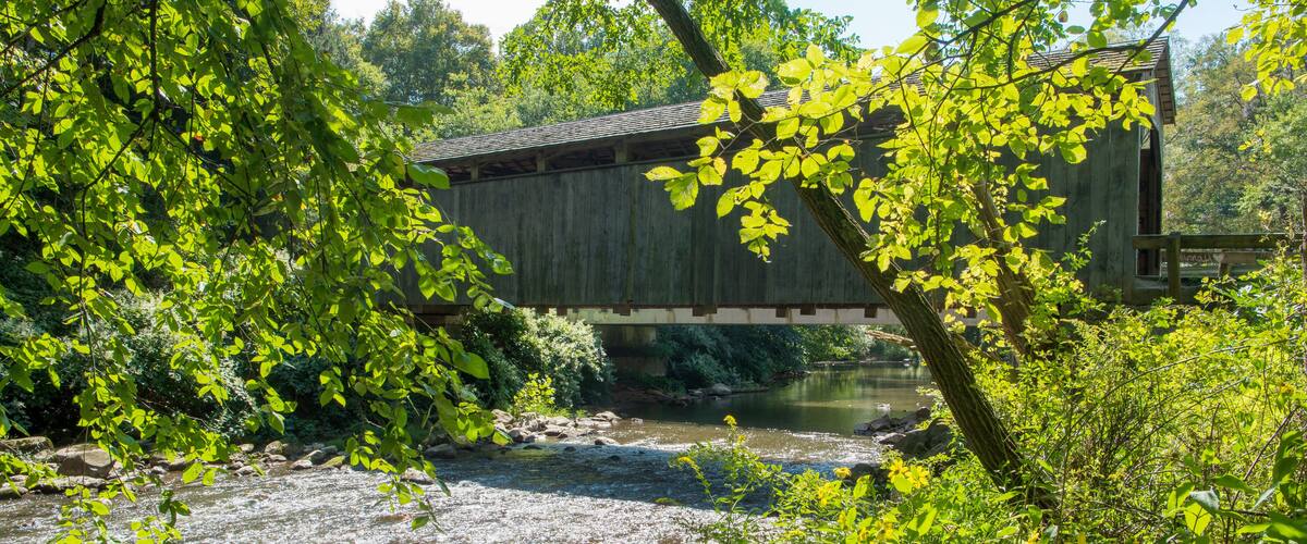 A wooden covered bridge over a river, surrounded by lush greenery and trees.