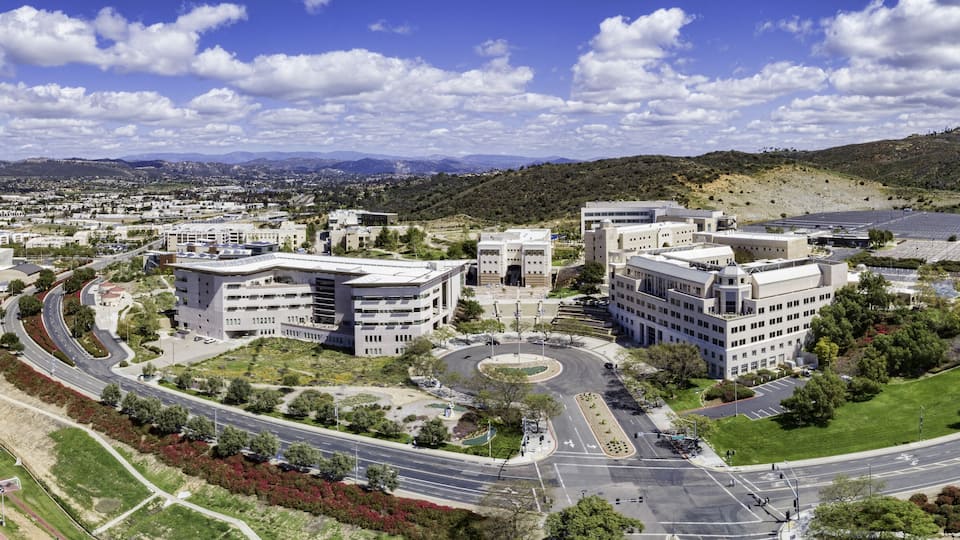 A modern urban area with a central roundabout, surrounded by multi-story buildings, green spaces, and a hilly landscape in the background.