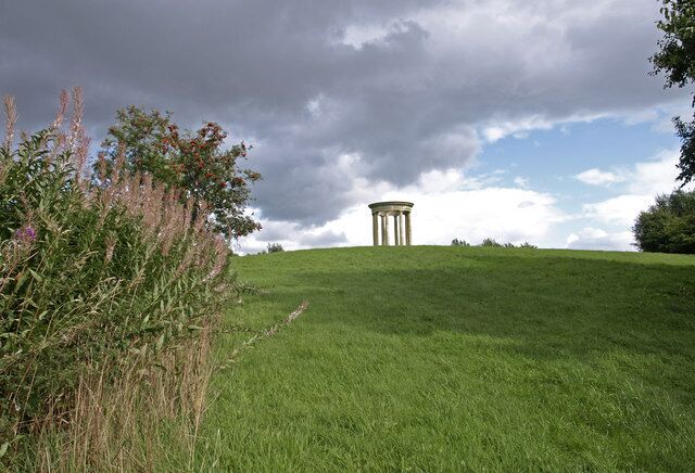 A grassy hill with a classical-style monument in the distance under a cloudy sky.