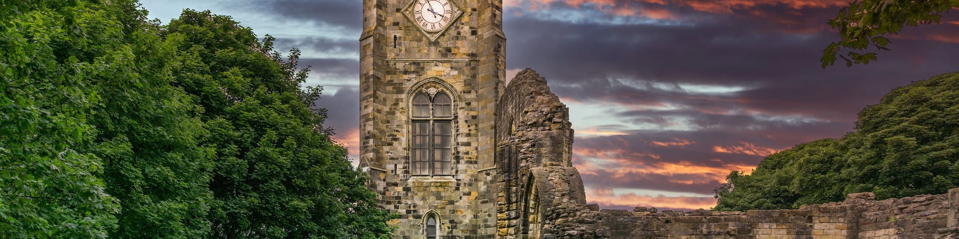 A historic stone tower with a clock, surrounded by trees and ruins.